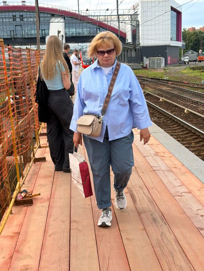 A blonde-haired woman wearing sunglasses, a blue striped button-down shirt, and grey pants walks on a wooden platform at a train station in Shcherbinka, Russia. She carries a beige crossbody bag and a white shopping bag with a red accent. To her left, a woman with long blonde hair, wearing a light blue top and striped pants, stands near an orange safety fence. Behind them, other individuals are visible. Train tracks run alongside the platform, and modern buildings, including a white structure with red accents and a red-arched building, are in the background. The scene appears to be outdoors during daylight.