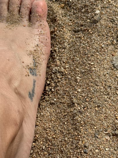 A close-up, ground-level shot shows a person's bare foot partially covered in sand. The foot is positioned on the left side of the frame, with the toes pointing towards the top left. Sand grains adhere to the skin, particularly on the top of the foot and between the toes. A faded black tattoo is visible on the side of the foot, appearing to be script. The background consists of a dense layer of small pebbles and sand, with a slight blur indicating a shallow depth of field. The lighting suggests it is daytime, with sunlight casting subtle shadows. The scene takes place on a beach or sandy area.