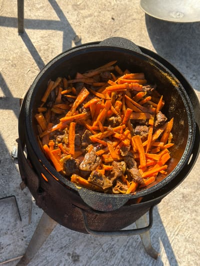 The image, likely taken outdoors in daylight in Большая Шильна, Russia, shows a cast iron pot, presumably a kazan, containing a dish of meat and julienned carrots. The pot is positioned on a stove or stand of similar material. The ground is concrete. A partial view of a lid sits to the left. The scene is still, with no apparent movement.