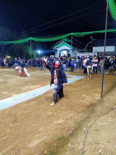 An outdoor nighttime scene in Tamansari, Indonesia, depicts a cultural performance on a dirt ground. The central figure, facing slightly left, wears a red-faced mask with dark, shaggy hair, a dark open jacket over a patterned top, and dark patterned lower garments, performing barefoot with their right arm raised. To the left, another blurry figure in motion appears to be performing. A large crowd of spectators stands in the background, observing the event. Behind the crowd, a light-colored building with a light green roof is visible under several artificial lights. Green netting or mesh is strung horizontally overhead, supported by poles, with power lines visible against the dark sky. A dark pole stands on the right, and a black cable crosses the foreground. Light blue lines are painted on the dirt ground, delineating a performance area.
