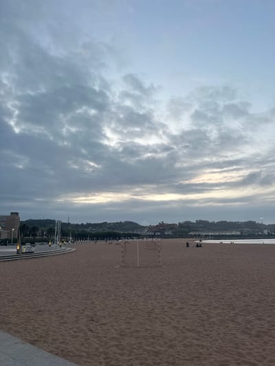 The image presents a wide view of San Lorenzo Beach in Gijón/Xixón, Spain, during dusk or dawn under an overcast sky. The foreground is dominated by a vast expanse of reddish-brown sand exhibiting subtle undulations. In the mid-left area of the beach, a white-framed portable soccer goal with a net stands on the sand. Further along the beach towards the right, multiple small groups of individuals are visible; one group of approximately three to four people is seated on the sand near the water's edge, adjacent to a dark, low object. A calm, dark blue-grey body of water (the sea) borders the beach on the right side.

A paved promenade runs parallel to the beach on the left, separated by a multi-tiered concrete structure. This promenade is lined with regularly spaced modern light poles emitting a warm glow, with some stacked white loungers positioned near the poles. Behind the promenade, a multi-story building with a light-colored facade is visible, partially obscured by palm trees. The background features a series of green, undulating hills covered in vegetation, interspersed with various urban buildings. Distant industrial structures or towers are faintly visible against the horizon line on the hills. The sky is predominantly covered by grey, textured clouds, with a brighter band of light visible near the horizon, suggesting the position of the sun. Patches of pale blue sky are discernible in the upper regions, and a faint white airplane contrail streaks across the upper right quadrant. Artificial lights from the promenade and distant structures are active, indicating low ambient light conditions.
