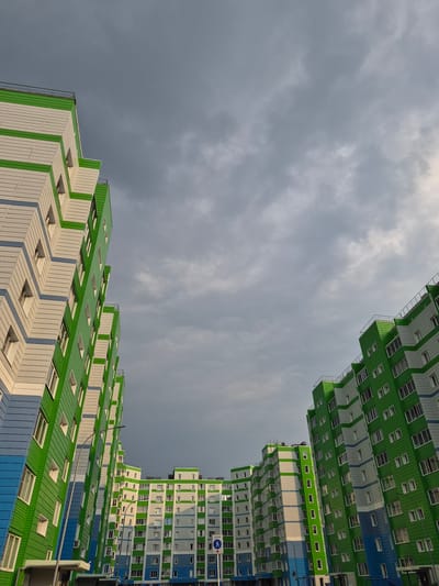 The image shows a low-angle shot of modern apartment buildings in Nyagan, Russia, against a cloudy sky. The buildings are tall and feature a distinctive architectural style with repeating geometric patterns in green, white, and blue. The facades are clad in panels, with numerous windows visible. The perspective is from below, looking upwards towards the sky, creating a sense of scale and vastness. The sky is filled with heavy, gray clouds, suggesting overcast weather, possibly before or after rain. The lighting is diffused, with some areas of the buildings catching subtle sunlight, hinting at a time around late afternoon or early morning. No people are visible in the image. There are no specific event cues, and the scene is static. One visible sign has Cyrillic text and an international symbol. The overall impression is one of contemporary urban architecture set against a dramatic, moody sky.
