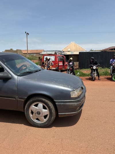 A dark grey sedan is parked on a dirt road, partially obscuring a scene with a red van and several individuals on motorcycles. The sedan's front left wheel and bumper are visible. The red van, labeled "VICTOR SCHOOL," is stopped, and a person wearing a reflective vest is standing next to it, looking forward. Three men are seated on motorcycles behind the van. The man on the leftmost motorcycle is wearing a black beanie and a black jacket. The man in the middle motorcycle is wearing a black beanie and a black outfit. The man on the rightmost motorcycle is wearing a purple shirt. The environment appears to be an urban setting in Nigeria, characterized by dusty roads, modest buildings in the background, and power lines overhead. The sky is clear and blue.