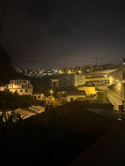 At night, the city of Câmara de Lobos, Portugal, is illuminated by streetlights and the lights from buildings situated on a hillside. The sky is overcast, contributing to the low-light conditions of the scene. Buildings of various sizes and styles populate the hillside, with visible rooftops and windows reflecting the city's artificial light. A crane stands out against the skyline. The city's infrastructure, including roads and power lines, is integrated into the environment. The darkness obscures detailed views of the vegetation and other natural elements of the landscape. A brick wall is on the edge of the frame.