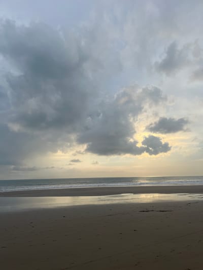 The image depicts a beach in Kerobokan Kelod, Indonesia under a cloudy sky at sunset. The beach occupies the right portion of the image, with visible wet sand and footprints. The ocean runs along the center, separating the beach from the sky. The sky is predominantly grey due to heavy cloud cover, with patches of sunlight breaking through.