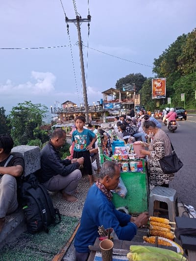 The scene takes place on a street corner in the Kecamatan Piyungan district of Yogyakarta, Indonesia. The coordinates are -7.846 latitude and 110.48 longitude. There are people on the sidewalk in front of a food stand. There are two men seated on the ground in front of the stand, and one man in a blue shirt, seated at a table. In the background there are a few people standing and walking and one man wearing a black shirt who is crouched down. They are all looking at the food stand. The stand has various products on display, and several individuals are seated around it. A young boy in a blue shirt stands to the left of the food stand. The stand is surrounded by a green metal frame. Next to the stand is a gray metal stool. The stand is covered in a black plastic tarp and a green cloth. The road is paved with asphalt. There is a tall power pole with wires running along it. There is a sign on the right of the image. The sign is orange with black lettering. Behind the power pole and the sign is a hillside with green trees growing on it. There is a building visible in the background to the left. The sky is blue with white clouds. The road is empty except for a few parked motorbikes and a car that is driving away. There are trees on the right side of the image, and a sidewalk on the left side. There is a concrete wall in the foreground, and a green mat on the ground.