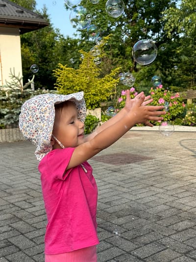 An outdoor, eye-level shot captures a young child, likely a toddler, enthusiastically playing with bubbles in Wisła, Poland. The child is positioned on the right side of the frame, reaching upwards with both hands towards numerous shimmering bubbles floating in the air. They are looking up with an open mouth, suggesting a joyful expression, possibly laughing or smiling.
The child is wearing a light-colored sun hat adorned with a small floral pattern, a bright pink short-sleeved t-shirt, and pink bottoms. Many bubbles of varying sizes are scattered across the frame, reflecting light and displaying iridescent, rainbow-like colors.
The background features lush green trees and bushes, some with clusters of pink flowers. The ground beneath the child is a paved surface made of grey, rectangular blocks arranged in a herringbone pattern. In the lower left background, a portion of a building with light-colored walls and a dark roof is visible. The bright lighting indicates it is daytime. No text is visible in the image.