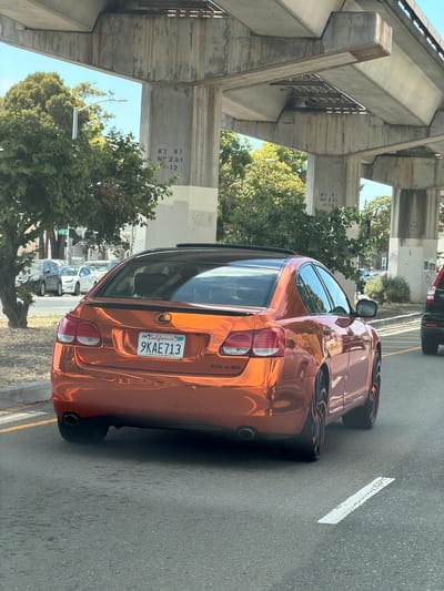This is a close-up, eye-level shot of the rear passenger side of a shiny orange Lexus GS 430 driving on a street in Oakland, California, USA. The car is in motion, indicated by the slight blur. The car has a California license plate that reads "9KAE713". The vehicle is being driven under an elevated highway structure with concrete pillars and overhead beams. The sky is visible through the structure, and there are trees and other cars in the background. The road surface is asphalt with a white line marker. The time of day appears to be daytime due to the lighting.