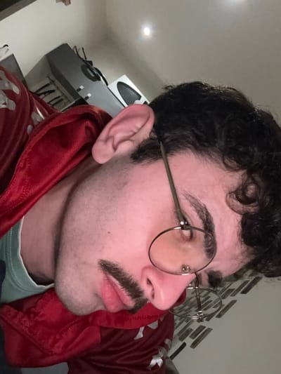A person with dark, curly hair, wearing round glasses, and sporting a mustache looks off to the side. The individual wears a red hooded jacket over a pale green shirt. Their skin tone is fair. Part of a kitchen appliance and some wall decorations are visible in the background.