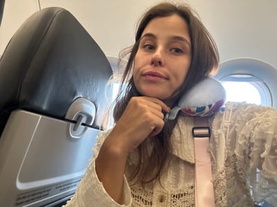 The image shows a woman seated on an airplane, likely in Incheon, South Korea, given the context of airplane travel. She is taking a selfie, resting her chin on her hand. A light blue neck pillow with a patterned design is visible around her neck. She is wearing a white lace top. The seat in front of her, which appears to be an airplane seatback, is visible on the left side of the frame, with text in Vietnamese and English that reads "Cài dây an toàn trong khi ngồi" and "Fasten seat belt while seated". The airplane window is visible on the right. The overall environment suggests travel.