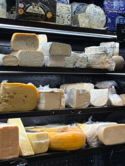 This close-up, slightly overhead photograph captures a diverse selection of cheeses displayed within a refrigerated case, likely in a market or specialty food store in Jerusalem, Israel. The cheeses, of various shapes including blocks, wedges, and partial wheels, are predominantly wrapped in clear plastic film, showcasing their different textures and hues, from creamy white to pale yellow and vibrant orange. Several blue cheeses are visible, some with distinctive veins of mold.
On the left side of the display, a dark blue and black package prominently features the brand "PAPILLON" and "ROQUEFORT," along with French text stating "AU LAIT CRU DE BREBIS" (from raw sheep's milk) and "AFFINÉ DANS NOS CAVES NATURELLES" (aged in our natural caves). A "SIGNATURE" label and a date "1906" are also discernible. Another partially visible label on a wrapped cheese contains the text "AWSOM," possibly part of a Wensleydale cheese brand. A small white label with "10" is seen above a blue cheese. Black labels attached to the display shelves show partial text such as "pl" and "122/008," possibly indicating prices or product codes, while other numbers like "35" and "15" are visible on the shelving itself. The display is well-lit, suggesting artificial lighting common in retail environments.