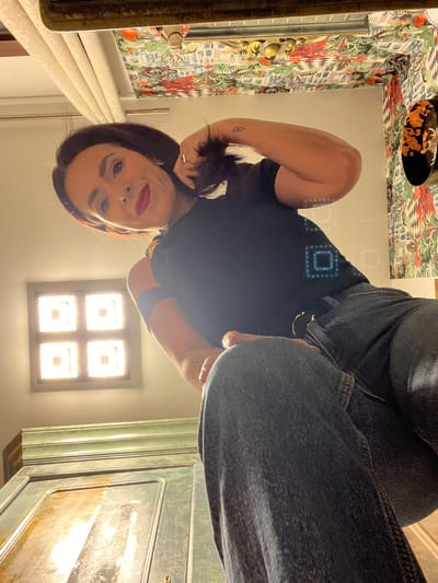 A woman with dark hair pulled back in a ponytail, wearing a dark t-shirt and jeans, is reflected in a mirror. She has on red lipstick and is wearing rings and earrings. The background features patterned wallpaper, a glass block window, and a pale green cabinet. The location is potentially a bathroom in Konz, Germany, but there is no information to confirm this.