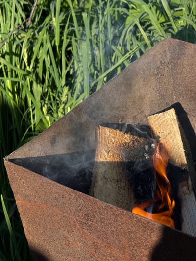 The image shows a rusty metal brazier angled diagonally in the frame. Inside, wood is burning with flames visible on the right and smoke rising. The brazier sits on green grass. The location is not specified in the image.