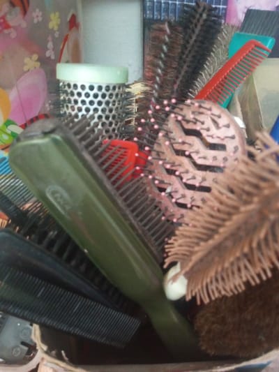 The image shows a collection of various hairbrushes and combs, clustered together in a container. There are round brushes, paddle brushes, and combs with varying bristle types and colors, including green, black, brown, and red. The brushes appear to be well-used and have a mix of plastic and natural bristles. The items are packed closely together, obscuring the details of the container.