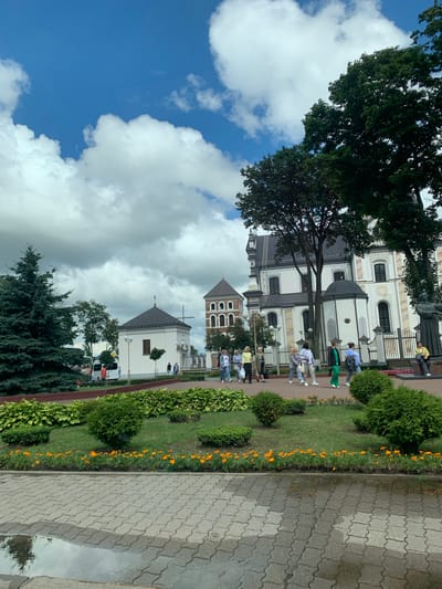 The image depicts the Niasvizh Castle complex in Niasvizh, Belarus, under a partly cloudy sky. In the foreground, a paved walkway with a puddle reflects the scene. A manicured garden with trimmed bushes and a line of small orange flowers separates the walkway from a grassy area. Several people are walking along a path in front of the castle complex. The castle itself is a large, white building with a dark roof, featuring numerous windows and architectural details. Adjacent to the main building is a smaller, white structure with a dark roof and a cross, and further to the left, a taller, brick tower with arched windows. Green trees, including a prominent evergreen on the left, are scattered throughout the scene. A statue of a man in a seated position is visible on the right side of the image. Several vehicles, including a white van, are parked in the background. The overall scene is that of a historical and cultural landmark with visitors enjoying the grounds.