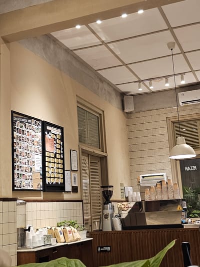 The image shows the interior of a cafe in Yogyakarta, Indonesia. The cafe has a minimalist design with light-colored walls, white tiled sections, and wooden accents. There are two framed displays on the wall, one with numerous small photos and another with gold-colored plaques. A coffee machine with a grinder is visible on the counter, along with stacks of disposable cups and several bags of coffee beans. Plants are placed on the counter, adding a touch of greenery to the space. Lighting fixtures are mounted on the ceiling and a pendant light hangs over the counter. A window with louvered blinds is visible on the right side of the image. A sign with text in Indonesian and a logo is also present on the wall. The overall impression is that of a clean and modern cafe.