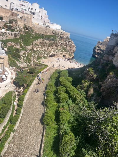 The image depicts a high-angle view of a beach and surrounding area in Polignano a Mare, Italy, on a bright day. The scene is dominated by a light-colored pebble-covered pathway running through the middle of the frame, flanked by vegetation and leading to a sandy beach.
On the left, there are white buildings constructed on the cliffs. Some people are on the pathway, seemingly walking towards the beach or lounging on the edges. The beach itself is populated with many people, suggesting a tourist or recreational area.
The Adriatic Sea extends in the background. The sky is a clear, gradient blue. On the right of the pathway is dense green foliage.