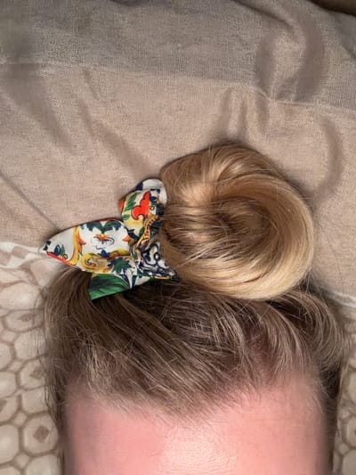 A close-up, top-down view depicts the upper portion of a person's head. The subject's hair, light brown at the roots, transitions to a lighter blonde shade, gathered into a coiled bun atop the head. This bun is secured by a scrunchie featuring a white background with a vibrant, multi-colored floral or abstract design in red, orange, yellow, blue, and green. The visible forehead and upper temples display a light skin tone with reddish undertones. The subject lies on a soft, textured, light beige fabric surface, possibly bedding, exhibiting folds and a fine weave. A portion of a second beige fabric, featuring a repeating geometric pattern, is visible to the lower left. No specific actions or interactions are depicted. The presence of the subject in the city of Catania, Italy, cannot be confirmed from the visual information provided within the image.