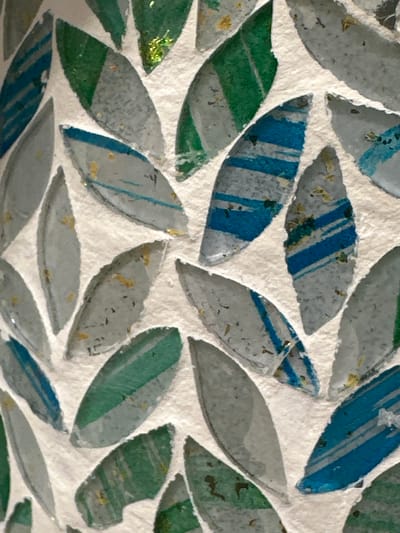 The image shows a close-up of a mosaic made of small, leaf-shaped pieces of glass and possibly other materials. The mosaic features a mix of colors, including various shades of green, blue, and grey, with some pieces having iridescent or metallic flecks. The background is a textured, light-colored material, likely the surface onto which the mosaic is applied. The arrangement of the pieces creates a pattern reminiscent of overlapping leaves. There are no people, animals, or discernible landmarks that would indicate a specific location in Coquitlam, Canada, or anywhere else.