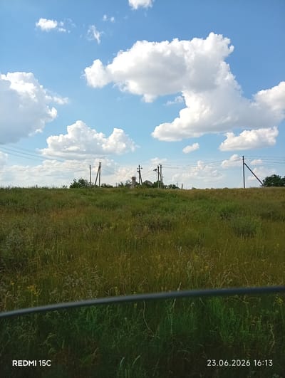 The image shows a wide, grassy field under a bright blue sky with fluffy white clouds. Several telephone poles with wires stretch across the horizon. In the distance, there appear to be a few houses and a small structure that might be a transformer. The foreground is dominated by tall, green and yellow grass. A dark, horizontal bar runs across the bottom of the image, obscuring a portion of the grass. The bottom right corner of the image displays the text "23.06.2026 16:13" and the bottom left corner displays "REDMI 15C". The scene suggests a rural or suburban landscape.