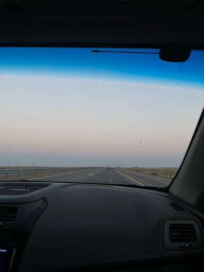 The image depicts a view from the interior of a vehicle traveling on a highway. The foreground shows the dashboard of the car, with a portion of the steering wheel visible on the left. The dashboard has a textured surface and features an air vent on the right side. A digital display on the lower left corner shows the time as 04:58.
The windshield of the car looks out onto a highway stretching into the distance. The road is paved with lane markings and a yellow line on the right shoulder. Guardrails line the left side of the highway. The environment appears to be a flat, open landscape with sparse vegetation and utility poles visible on the horizon. The sky transitions from a clear blue at the top to a soft gradient of pink and light purple, suggesting either dawn or dusk. The sun is not directly visible. There are a few distant vehicles on the highway. The scene is set on a highway in the vicinity of Тоқсансай, Kazakhstan.