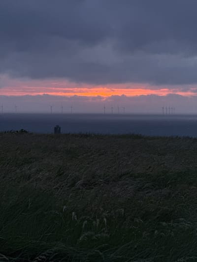 The image displays a dramatic landscape scene of an offshore wind farm, consistent with the location context of Fécamp, France, captured during either sunrise or sunset.
In the foreground, a dark expanse of tall, dense grass occupies the bottom right section of the image, suggesting a coastal vantage point, possibly a dune or cliff edge. Beyond this natural barrier, a vast body of water, likely the sea, stretches towards the horizon.
Across the mid-ground, numerous offshore wind turbines are visible, silhouetted against the radiant sky. These industrial structures stand tall and slender, their blades faintly discernible, forming an extensive line across the water. Towards the center of the image, a more prominent, dark, rectangular structure, possibly an offshore substation or platform, breaks the uniform line of turbines.
The most striking feature is the horizon, where an intense band of vivid orange and pink light glows brilliantly, illuminating the scene and indicating the presence of the setting or rising sun. This warm light contrasts sharply with the cooler, darker tones of the sky above, which ranges from deep blues and greys in the upper left, transitioning to lighter hues where it catches the sun's glow. Dramatic clouds are scattered across the sky, partially illuminated by the powerful light source.
The scene conveys a serene yet dramatic atmosphere, highlighting the intersection of natural beauty and industrial innovation. No people or visible text are present in the media file.