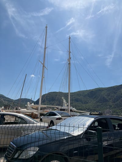 The image shows a view from Kotor, Montenegro, featuring several tall sailing ship masts in the harbor. Lush green mountains rise in the background. Parked in the foreground, behind a wire fence, are multiple cars, including a taxi. The sky is blue with scattered clouds.
