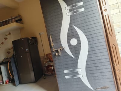 An indoor, slightly angled shot captures a modern door with a grey and white abstract pattern dominating the right side of the frame. The door features a metallic handle and latch. To the left, a domestic interior is visible, likely a kitchen or utility area. A dark-colored double-door refrigerator stands prominently in the center-left. Above it, a pale yellow wall features decorative floral decals, and a wall-mounted shelf holds several jars, likely containing spices or food items. An electrical outlet with a plugged cord is on the adjacent yellow wall. On the floor, near the door, a small, ornate wooden Pooja Mandir (shrine) containing religious figures is present. An orange hose or cable is also on the floor. The setting suggests a home in Dhandhuka, India, under ambient lighting. No individuals are present, and no specific activity is taking place. Text on the refrigerator and jars is illegible.