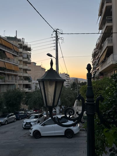 The image captures an urban street scene in Athens, Greece, during twilight. The foreground is dominated by a decorative black lamppost with ornate scrollwork, partially obscuring a white Volkswagen Polo. In the midground, several cars are parked along the street, including a silver hatchback, a dark blue sedan, and a white Lexus. The street curves gently uphill, leading the eye towards the background where apartment buildings line both sides. These buildings feature balconies with awnings and are interspersed with trees. Power lines crisscross the sky, and a utility pole is visible. The sky displays a gradient of pale blue to soft orange, indicating the setting sun, with a faint silhouette of hills on the horizon. The overall impression is a residential street in Athens as daylight fades.