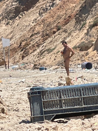 The image depicts a man standing nude on a beach, holding an orange object in his right hand. He is facing to the left of the frame and his left hand is on his hip. The background consists of a rocky hillside and a beach littered with various debris, including a large black barrel and what appears to be a discarded metal cage in the foreground. A white sign on a pole is visible to the left of the man. The location is the coast of Ga'ash, Israel.