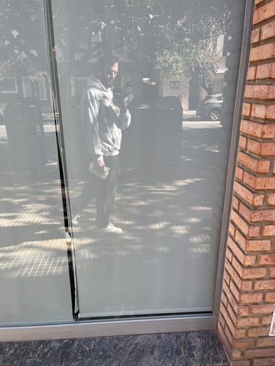 A reflection of a person seated on a curb, and the surrounding urban environment, is visible on a closed window screen. The person appears to be wearing a light-colored hooded sweatshirt and dark pants. Trees, a building, and a car are also part of the reflection. The window frame is set in a building constructed of brick and a type of grey stone. The location is potentially in Cordoba, Argentina.