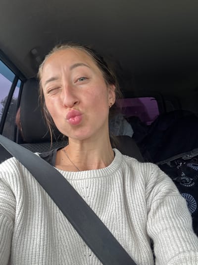 An adult female is depicted in the driver's seat of a vehicle, taking a selfie-style photograph. She has fair skin with visible freckling across her cheeks and nose, and light brown hair is partially pulled back. Her left eye is closed in a wink, and her lips are pursed into a "kiss" expression. Her visible right eye has a light blue-green iris. She wears a light cream-colored, ribbed knit sweater over a dark undershirt, and a thin silver-colored necklace is visible around her neck. A small gold stud earring is present in her right ear. A dark gray seatbelt crosses diagonally from her left shoulder to her right hip, indicating it is buckled. The car's interior features a dark gray headliner and dark upholstered seats, with a headrest directly behind her. Through the driver's side window, a blurred outdoor scene reveals green foliage and a muted sky, suggesting an overcast or twilight setting. The rear window of the vehicle exhibits a distinct purple tint, and various items, including dark fabric and a black and white patterned bag, are visible in the back passenger or cargo area.
