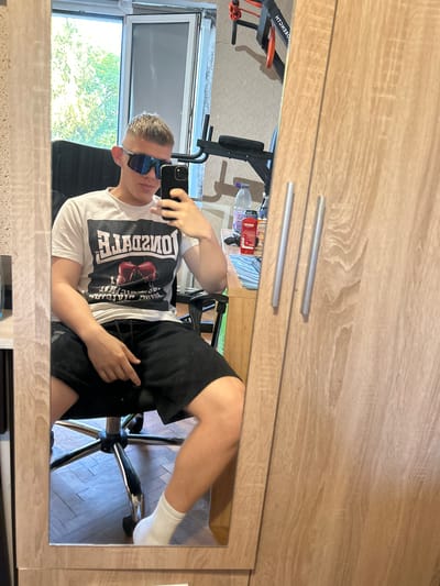 A person is reflected in a mirror. The individual is wearing a white t-shirt, black shorts, and white socks. They are seated on a chair. The background includes a window, trees, and exercise equipment. The location cannot be determined to be the city of Galabovo, Bulgaria with the available information.