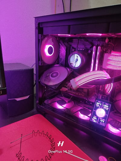 A black computer case with a glass side panel is lit up with pink and white LED lights. The case is on a red mat. Inside the case are various components, including a graphics card, a motherboard, and several fans. The graphics card is a GEFORCE RTX, which is illuminated by pink LED lights. The fans are also illuminated by pink LED lights, and one fan is illuminated with white LED lights. A monitor with a digital display is in the background. The digital display shows system information, including CPU temperature, RAM usage, and network usage. The coordinates provided are located in the city of Rajshahi, Bangladesh. There is a black speaker to the left of the case. On the red mat, a thin black wire in the shape of a coil can be seen.