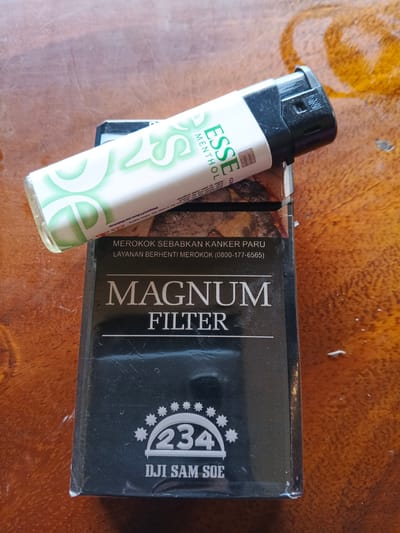 The image shows a pack of Magnum Filter cigarettes with an Esse Menthol cigarette pack placed on top of it. A black lighter is resting on top of the Esse Menthol pack. The items are placed on a wooden surface, possibly a table. The background is a close-up shot of the wooden surface. There is no indication of the location in the city of Geulumpangsulu Timu, Indonesia.