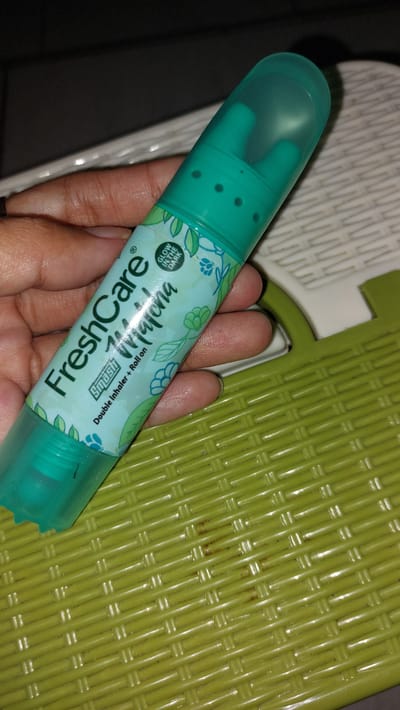 A hand is holding a teal-colored "FreshCare" product, which appears to be a double inhaler and roll-on. The product has a clear plastic cap that covers two distinct applicator ends. One end, visible under the cap, has a few small holes, suggesting it might be an inhaler. The other end, visible at the bottom, has a rollerball mechanism, indicating it's a roll-on.
The body of the FreshCare product is decorated with a light blue and green floral and leaf pattern. Some text is visible on the product. In a circular logo with a white background and black text, it reads "GLOW IN THE DARK". Below that, in large, stylized black font, are the words "FreshCare" and "Yuina". Smaller text to the right of "FreshCare" states "Double Inhaler + Roll-on". There is also a small registered trademark symbol.
The hand holding the product has light to medium skin tone and is illuminated by what appears to be artificial light, given the warm tones and shadows. The background is a textured, bright green surface, possibly a mat or a woven plastic material. To the left of the green surface, there's a glimpse of a white patterned surface and a dark area, possibly indicating an indoor setting. The overall impression is a close-up shot of a personal care item being presented. The image suggests a moment of everyday use, with no specific time of day or weather conditions discernible. The context is identified as Karangjati, Indonesia.