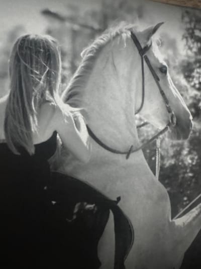This black and white, grainy photograph captures an intimate interaction between a person and a horse, presented with a 90-degree clockwise rotation.

The primary subjects are a light-colored horse, likely white or grey, and a person whose back and long, light-colored hair are visible. The horse, equipped with a bridle and a dark saddle, has its head raised, looking towards the top of the frame. Its mane is full, and highlights on its coat suggest a textured surface.

The person is seen from behind, positioned towards the bottom-right of the frame, with their head gently bowed. Their long, flowing hair cascades over their back and shoulder. They appear to be wearing dark clothing that merges with the saddle. One arm is extended towards the horse's neck or shoulder, suggesting a gesture of affection or connection.

The scene takes place outdoors, indicated by the blurred background of trees and foliage, hinting at a natural setting such as a field or forest edge. The lighting suggests it is daytime. The overall mood conveyed is one of calm, tenderness, and a strong bond between the human and the animal. No visible text is present in the image. The location context provided is Wattrelos, France.