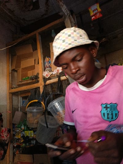 An indoor scene in Gama, Nigeria, depicts a young adult male with dark skin, wearing a checkerboard-patterned bucket hat and a pink short-sleeved t-shirt with a Nike swoosh and an FC Barcelona crest. He is observed looking downwards, holding a dark-colored mobile device in his left hand and a purple-capped stylus or pen in his right hand, actively engaging with the device. The background reveals a cluttered retail environment, featuring a wooden and mesh shelving unit stocked with flattened cardboard boxes and various packaged goods. Several transparent plastic buckets, some containing what appears to be nuts or similar items, are also visible. Two red plastic bags hang from an overhead structure in the upper right quadrant of the image.