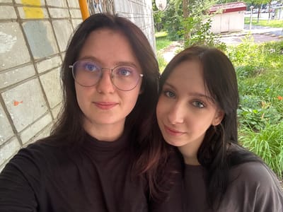 Two young women are taking a selfie outdoors. The woman on the left is wearing glasses and a black t-shirt. The woman on the right is wearing a grey t-shirt. They are standing in front of a brick wall with some yellow and grey paint on it. In the background, there are trees and a small building. The image was taken in the city of Obninsk, Russia.