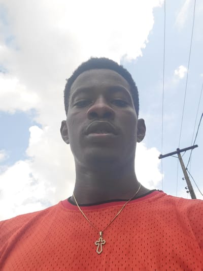 The image is a low-angle, close-up shot of a young Black male, taken outdoors against a bright, cloudy sky. The subject is wearing a bright orange mesh t-shirt and a gold chain with a cross pendant. His hair is short and dark, and he is looking directly at the camera with a relaxed expression.
The sky is mostly clear with scattered white clouds, suggesting it is daytime, likely midday due to the brightness. A utility pole with wires is visible in the background, indicating an urban or suburban setting. The image appears to be a selfie, given the perspective.
No text is visible in the image. The scene is casual and portrays a moment of personal documentation. The image has been rotated 90 degrees counter-clockwise.
