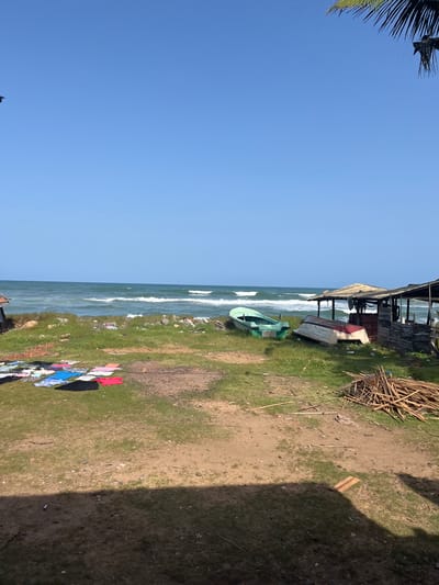 The image depicts a coastal scene in Moratuwa, Sri Lanka. The foreground shows a patch of dry, grassy ground with some scattered debris and a pile of wooden sticks. To the left, a collection of brightly colored laundry is laid out on the grass to dry.

In the middle ground, a small embankment separates the land from the ocean. Two small boats are visible on the grass near the embankment. One is green and upright, while the other is inverted and appears to be white with a red trim.

Further to the right, there is a rustic, open-sided structure with a corrugated metal roof, suggesting a small shelter or vendor stall. The sea stretches out to the horizon under a clear blue sky. Gentle waves are breaking on the shore, creating a serene coastal atmosphere. The image is taken during daylight hours, likely mid-morning or afternoon, given the angle of the sunlight casting shadows. There are no people visible in the frame.