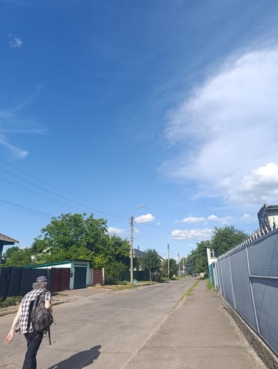 The image depicts a person walking away from the camera on a paved street in a suburban area. The person is wearing a checkered shirt, dark pants, and a backpack. They have their left arm extended as if gesturing or balancing. The street is lined with houses, fences, and trees. There are power lines overhead. The sky is a clear blue with scattered white clouds, suggesting a sunny day. The scene appears to be in a residential neighborhood, possibly a village or a less developed part of a town, given the presence of garages and older style fencing. The lighting suggests it is daytime, likely late morning or early afternoon due to the sharp shadows cast by the person and overhead structures. No visible text is present in the image. The overall mood is casual and everyday, with no indication of a specific event or strong emotion. The context provided indicates the location is Chernihiv, Ukraine.