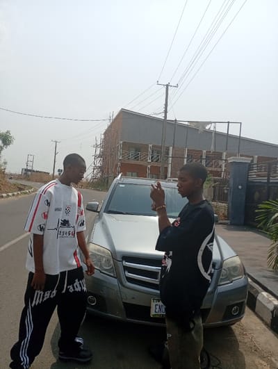 Two young men stand next to a silver Mercedes-Benz SUV. The man on the left wears a white T-shirt and black pants with white stripes and the words "mad lady" on the legs. The man on the right wears a black T-shirt and khaki pants. They are positioned on a paved road near a curb. In the background is a partly-constructed gray building, power lines, and trees. The sky is overcast.