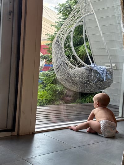 A baby in a diaper is crawling on a gray tiled floor in what appears to be an indoor setting next to a sliding glass door. Outside the door, there's a hanging white woven chair with a gray cushion and blue polka-dot ties, along with trees and parts of a building. The bottom of the door frame is visible in the foreground. The location is unknown.