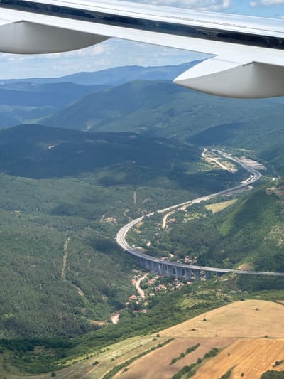An aerial view captures a highway with a viaduct passing through a valley in the vicinity of Stolnik, Bulgaria. The highway is curved and ascends the valley, with the viaduct supporting it as it crosses a lower section of the terrain. Numerous vehicles are visible on the highway and the viaduct. On either side of the highway, the landscape is covered with dense green forests and rolling hills. Small clusters of red-roofed houses are scattered throughout the valley, nestled among the trees and along the winding roads that lead to the highway. In the foreground, patches of dry, yellow fields indicate agricultural land. The sky is partly cloudy, with the wing of an aircraft visible in the upper portion of the image, suggesting the photograph was taken from a plane.