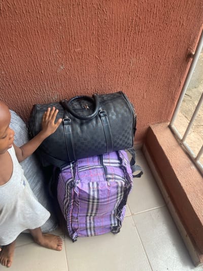 The scene is located in Oba-Aboji, Nigeria. A young child, possibly male, wearing a light-colored sleeveless top with a star graphic, stands barefoot on light-tiled flooring. The child's right hand is placed flat on a black duffel bag with a dark checkered pattern, equipped with top handles and a zipper. This duffel bag rests on a larger, purple plaid-patterned backpack made of woven material, which shows visible wear and fraying. Both bags are positioned against a reddish-brown, heavily textured wall. A light-colored metal railing with vertical bars is visible on the right, and a portion of a grey, fabric-covered object is present to the child's left.