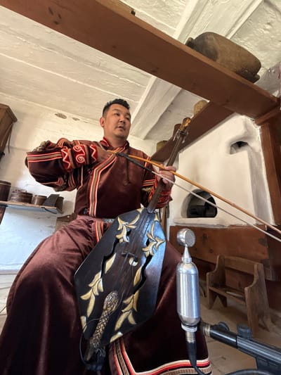 An East Asian man is seated in traditional clothing, playing a stringed instrument. The man is wearing a dark brown, long-sleeved robe with intricate red and yellow patterns on the cuffs and along the front opening. He is holding a bow, which is positioned across the strings of a wooden instrument that rests on his lap. The instrument has a dark, intricately carved body with gold leaf accents and a snake-like motif. In front of the man, a vintage microphone is attached to a stand. The background suggests an indoor setting with a white plastered wall, a white ceiling with exposed wooden beams, and rustic wooden furniture. There is a white stove or hearth visible in the background. The overall scene appears to be within a museum or historical exhibit. This image was taken in the Shushenskoye Museum of Folk Life and Crafts, located in Shushenskoye, Russia.