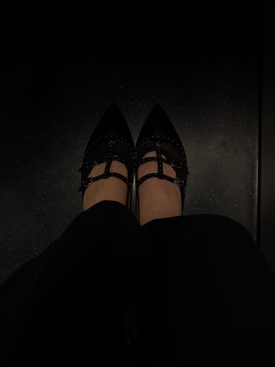 This overhead shot, taken in Glattbrugg, Switzerland, captures a person's lower legs and feet. The subject is wearing a pair of distinctive black, pointed-toe flat shoes, adorned with numerous silver studs around the edges and on multiple straps across the instep. These straps are secured with small, gold-colored buckles. The person's bare skin is visible on the top of their feet between the straps. The shoes are paired with dark, possibly black, clothing that covers the upper part of the legs, visible as a dark fabric mass on the right side of the frame.

The person is standing on a dark, speckled floor, which possesses a highly reflective, mirror-like surface. This surface creates a clear and sharp reflection of the shoes and lower legs directly beneath them, giving the impression of two symmetrical pairs of feet meeting at the toes. The lighting is dim, emphasizing the shoes and the immediate area of the floor, while the surrounding environment remains largely obscured in darkness. The speckled pattern of the floor is apparent both on the actual surface and in its reflection. No other people, objects, or specific activities are discernible, and there is no visible text. The indoor setting and artificial lighting prevent determination of the time of day or weather.