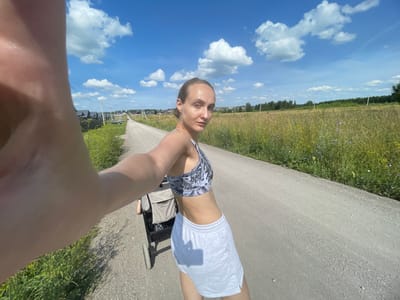 A woman is taking a selfie with a baby stroller on a gravel road in a rural area of Podshivalovo, Russia. The woman is wearing a sports bra and shorts. She has light brown hair pulled back in a bun. The baby stroller is light brown and has large wheels. The road is lined with fields of tall grass and wildflowers. In the background, there are trees and houses. The sky is blue with white clouds.