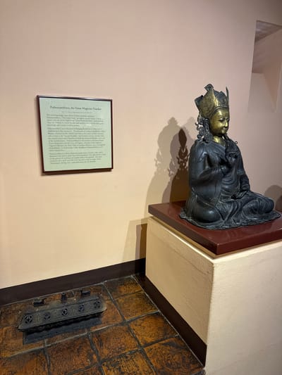 A medium shot depicts a religious artifact, Padmasambhava, the Great Magician-Teacher, displayed on a reddish-brown shelf in what appears to be a museum or gallery. The sculpture is predominantly dark with golden accents on the face and headgear. To the left of the sculpture, a framed description of Padmasambhava provides detailed information about the artifact, including the fact that it is a 17th-century copper sculpture. Below the shelf is what appears to be a tiled structure with an ornate black metal grate. The walls are a pale tan color. Based on the OCR text, the statue's origin is related to the Kathmandu Valley of Nepal. While not explicitly stated, Lalitpur, Nepal, is a part of Kathmandu Valley.